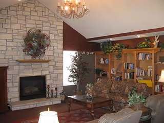 A cozy living room with a stone fireplace, a chandelier, a floral-patterned sofa, armchairs, a wooden coffee table, bookshelves filled with books and decorative plants, and a window letting in natural light.