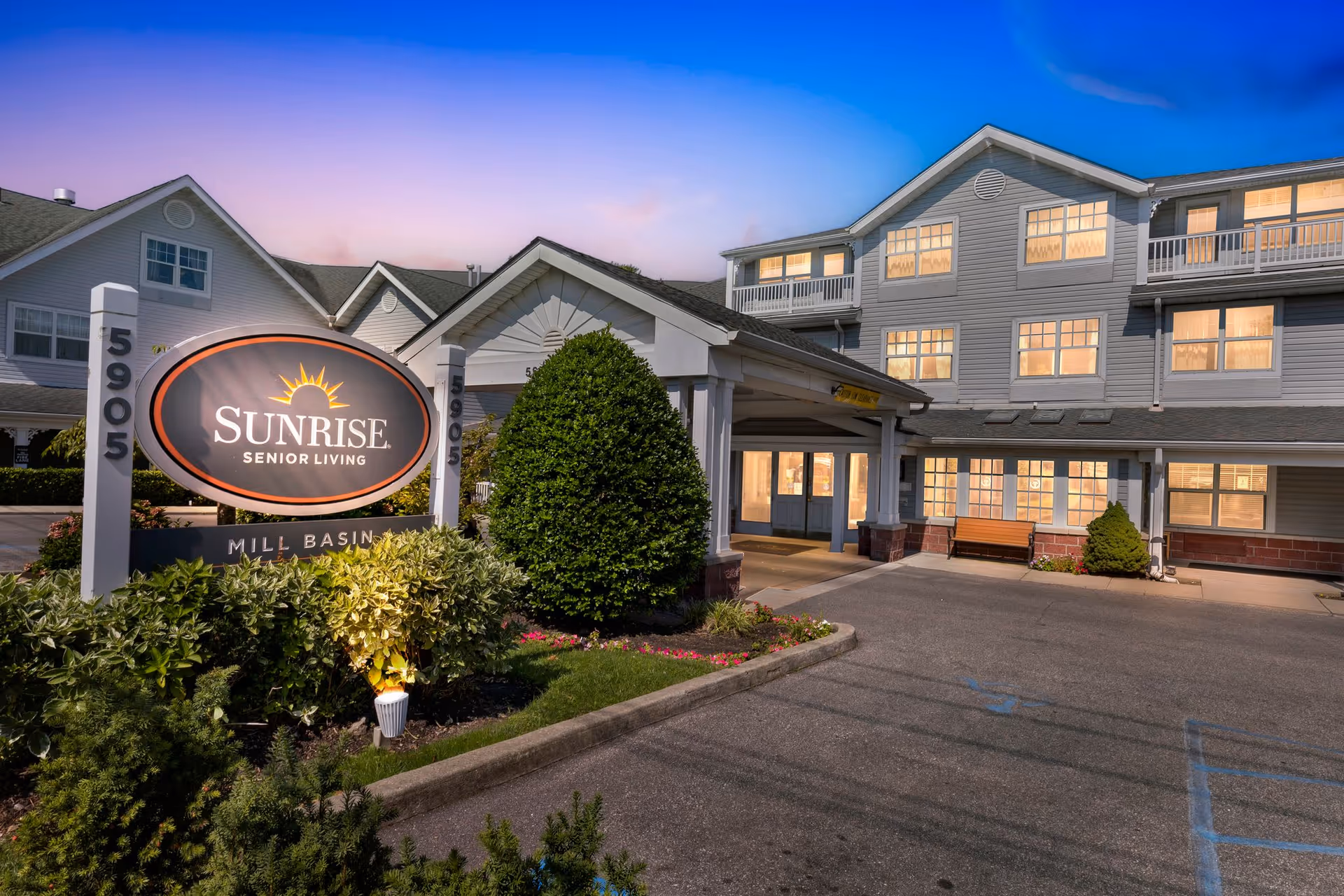 Exterior view of Sunrise Senior Living at Mill Basin during dusk, showing a multi-story building with many lit windows, a covered entrance, landscaped bushes, and a large sign with the facility name and address 5905.