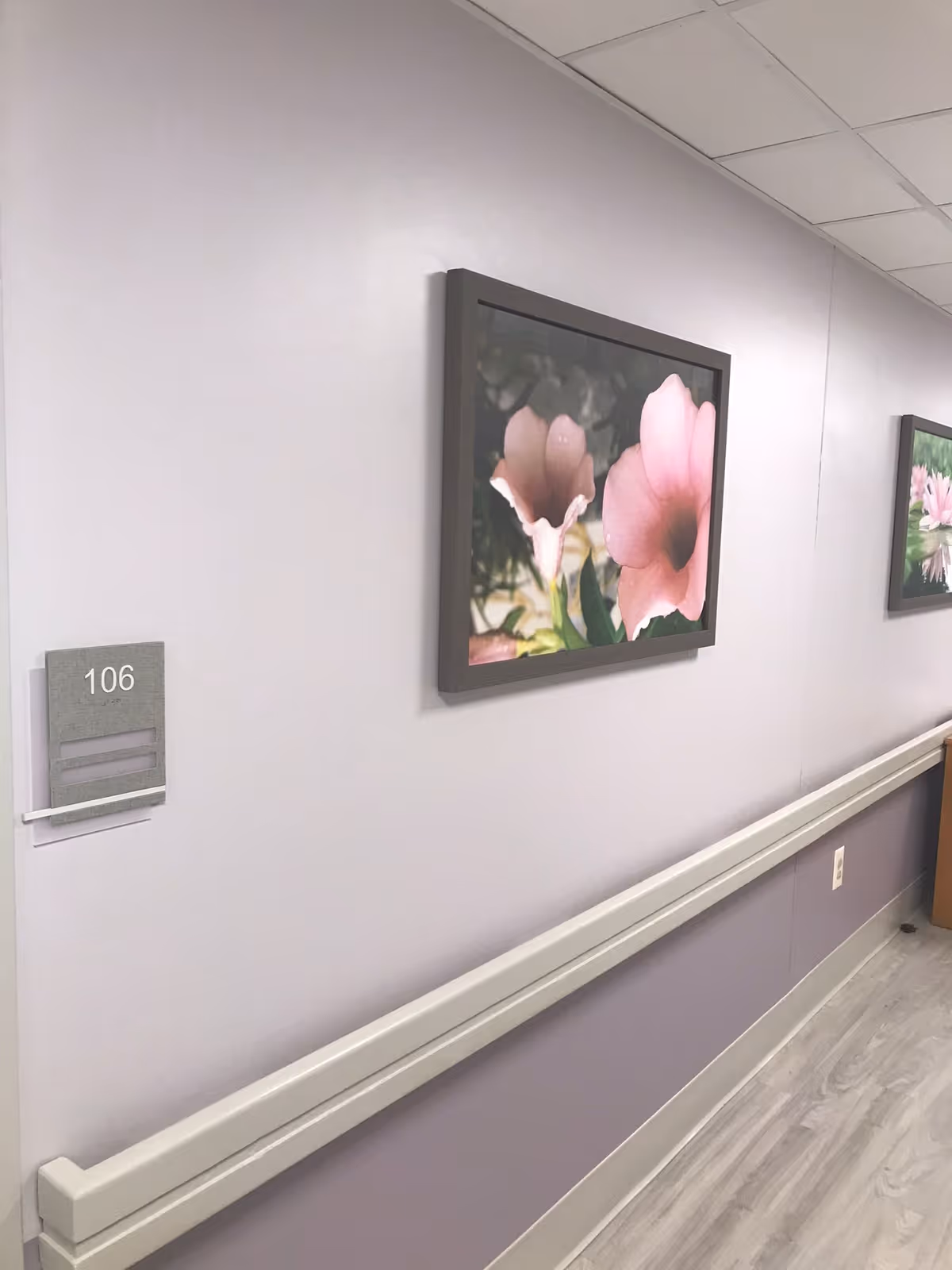 A hallway in a senior living facility with light purple walls and a handrail along the wall. There are framed pictures of pink flowers hanging on the wall, and a room number sign labeled 106 is visible.