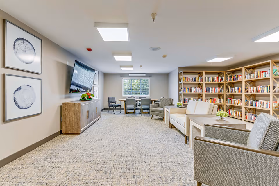 A bright and spacious common area in an assisted living facility featuring a large bookshelf filled with books, several armchairs and sofas arranged around small tables with flower vases, a flat-screen TV mounted on the wall above a wooden cabinet, and a table with chairs near a window with blinds.