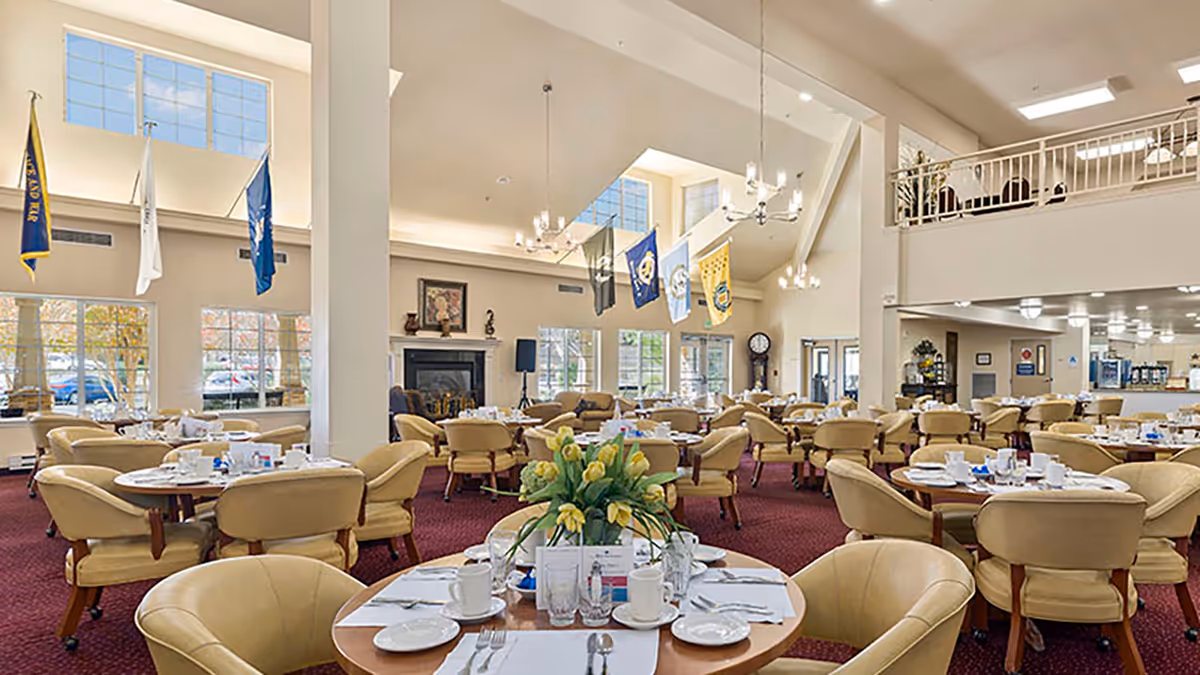 Large, airy dining room with round tables set for meals, beige chairs, high ceilings and flags hanging from above.