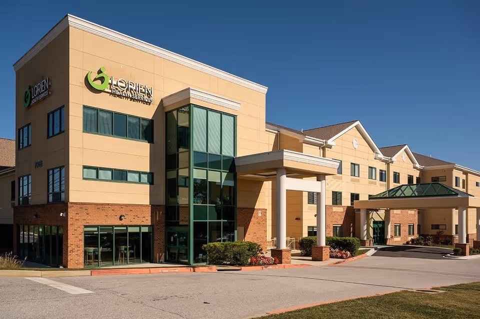 Exterior view of Lorien Elkridge Health Services building with a clear blue sky. The building features a combination of beige and brick facade with large green-tinted windows and a covered entrance supported by white columns.