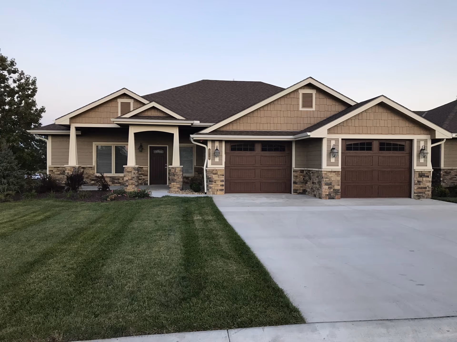 Front exterior view of a single-story residential building with a well-maintained lawn, two brown garage doors, a covered porch with stone pillars, and beige siding with stone accents.