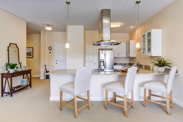Modern kitchen area with a large island countertop featuring three cushioned bar stools. The kitchen includes stainless steel appliances such as a refrigerator and a range hood. Pendant lights hang above the island, and white cabinets line the walls. To the left, there is a wooden console table with a mirror, plants, and decorative items.