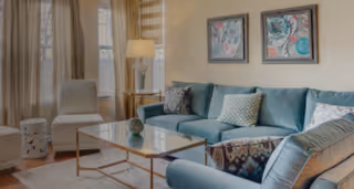 A cozy living room with a light blue sectional sofa adorned with patterned throw pillows, a glass coffee table with a decorative item, two white armchairs, a floor lamp, and framed artwork on the wall. Large windows with beige curtains allow natural light to fill the space.