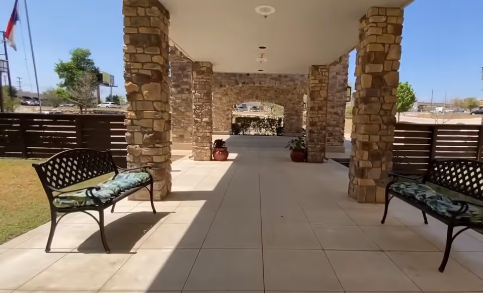 Outdoor covered walkway with stone pillars and tiled floor, featuring two black metal benches with cushions on either side. There are potted plants near the far end and a wooden fence enclosing the area. The sky is clear and blue.