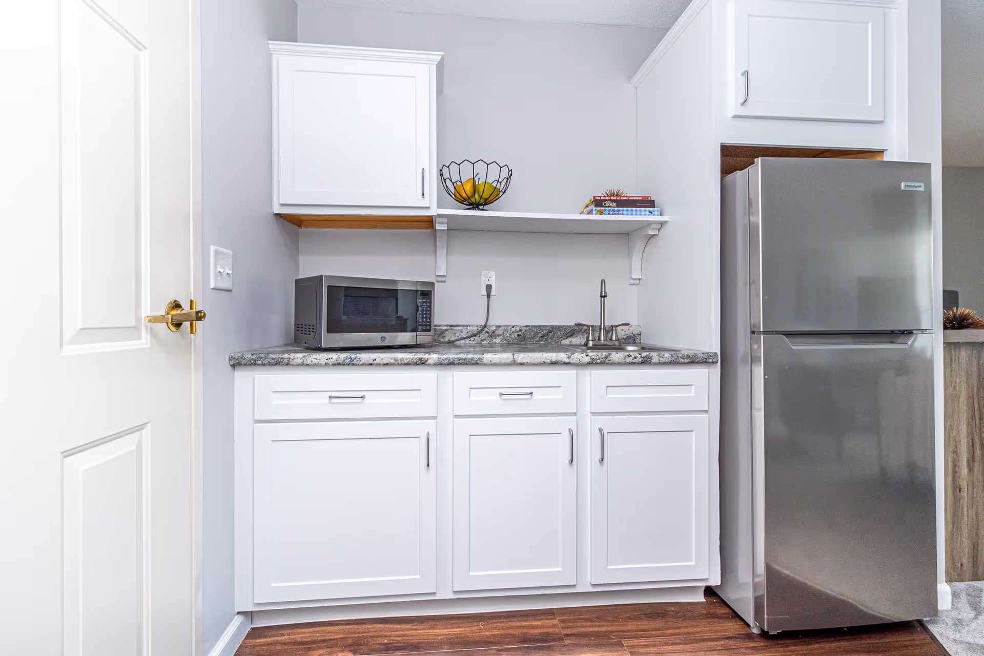 Small kitchenette with white cabinets, marble countertop, microwave, sink, fruit bowl on a shelf and a stainless steel refrigerator.