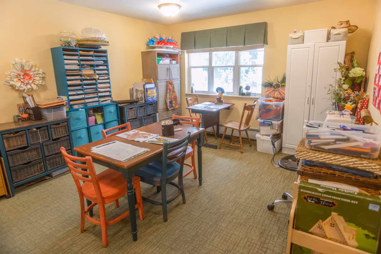 A well-lit activity room with a table and several chairs, surrounded by shelves and storage units filled with craft supplies and materials. The room has a window with a green valance, and various decorations and organized bins are visible throughout.