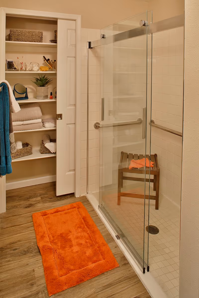 A bathroom with a glass-enclosed shower featuring a wooden bench inside. There is an orange bath mat on the wooden floor outside the shower. To the left, an open closet with shelves holds towels, baskets, toiletries, and a small plant. A blue towel hangs on a hook on the wall.
