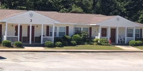 Single-story apartment building with white siding and brown doors, numbered 3 and 4, surrounded by green bushes and trees in the background, with a paved parking area in front.