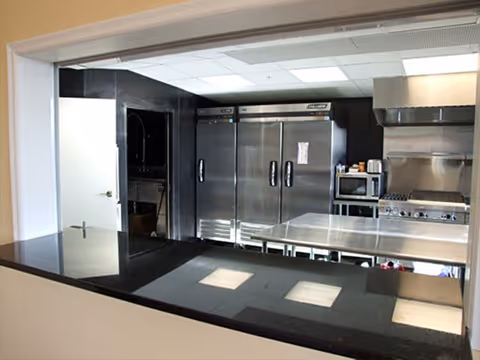 View through a pass-through window into a commercial kitchen featuring stainless steel appliances including a large refrigerator, microwave, stove with hood, and a stainless steel work table.