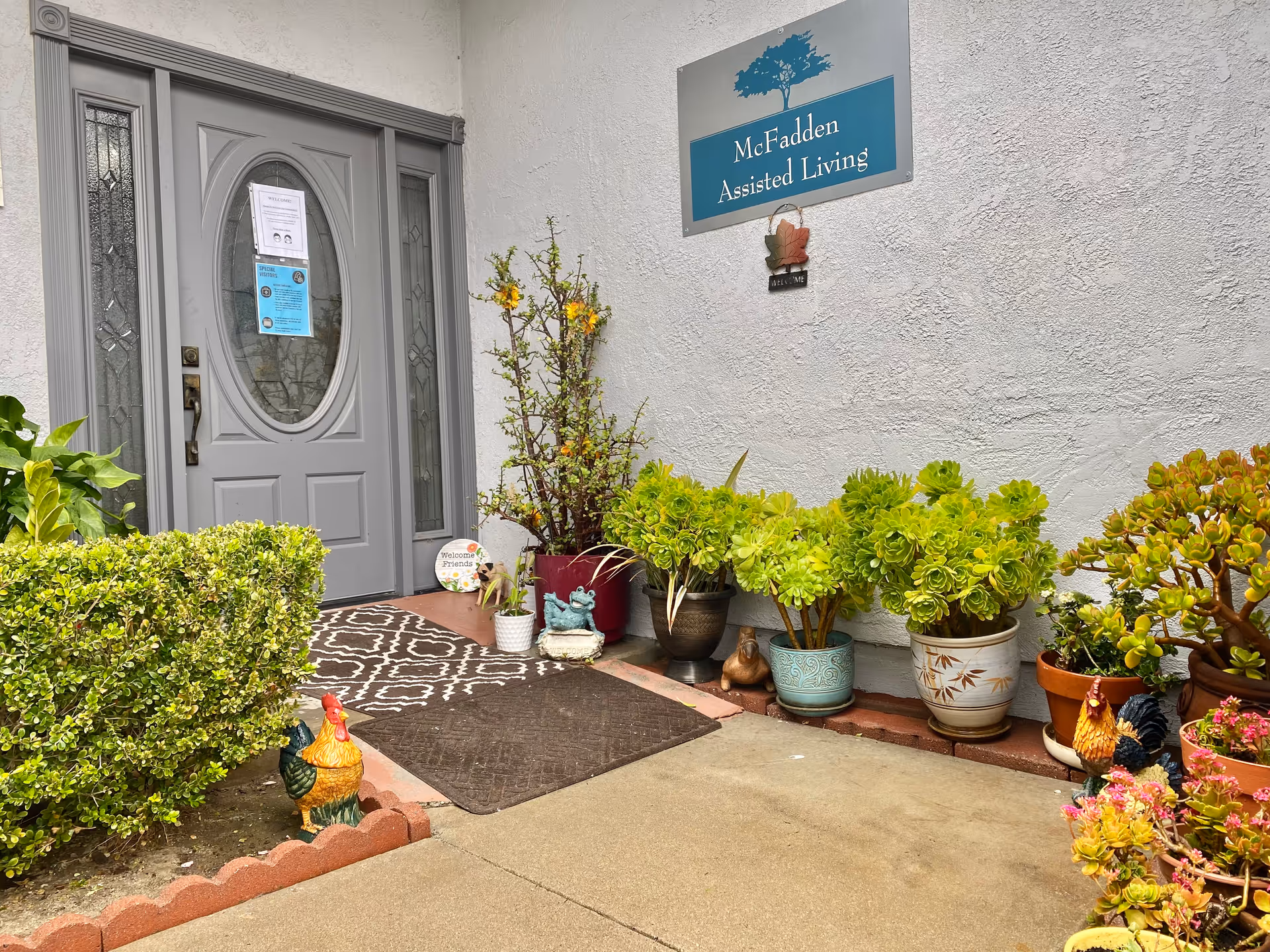 Entrance to McFadden Assisted Living with a gray door featuring decorative glass panels. The entrance area is decorated with various potted plants and small garden ornaments, including two rooster statues. A welcome sign and a McFadden Assisted Living sign with a tree logo are visible on the white exterior wall.