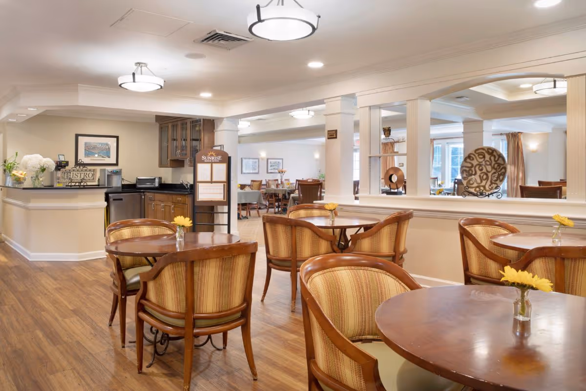 A bright and inviting dining area in a senior living facility with round wooden tables and cushioned chairs. Each table has a small vase with a yellow flower. The room features wood flooring, soft lighting from ceiling fixtures, and a counter area with cabinets and a coffee machine. Decorative plates and framed artwork adorn the walls, and a sign with the Sunrise logo is visible near the counter.