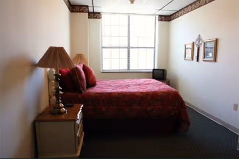 A simple bedroom with a bed covered in a red patterned bedspread and matching pillows. There is a wooden nightstand with a lamp on it next to the bed. The room has a large window letting in natural light, two framed pictures on the wall, and a small television in the corner.