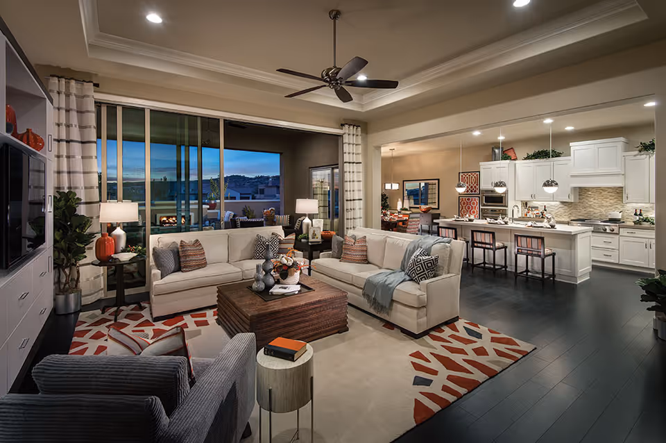A spacious and modern open-concept living room and kitchen area in a senior living facility. The living room features two beige sofas with patterned throw pillows, a wooden coffee table, a patterned area rug, and a ceiling fan. Large sliding glass doors open to an outdoor patio with seating and a fireplace, visible in the background. The kitchen has white cabinetry, a large island with bar stools, pendant lighting, and stainless steel appliances. The dining area is visible beyond the kitchen with a table and chairs.