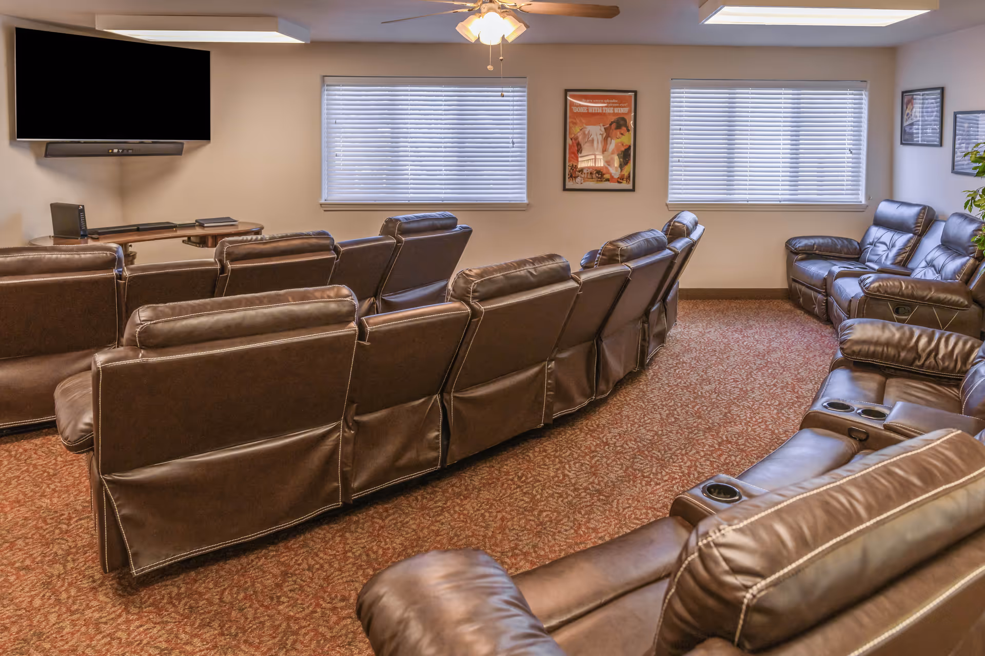 A cozy media room with multiple rows of brown leather recliner chairs facing a wall-mounted flat-screen TV. The room has two windows with blinds, framed movie posters on the walls, a ceiling fan with lights, and a carpeted floor with a patterned design.