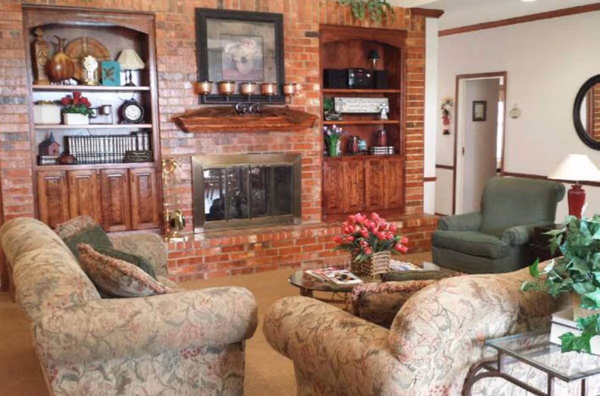 A cozy living room with floral patterned sofas and a green armchair arranged around a glass coffee table with a basket of red flowers. The room features a brick fireplace with a wooden mantel, flanked by built-in wooden shelves filled with decorative items, books, and a clock. A table lamp and a round mirror are visible on the right side, with a doorway leading to another room.