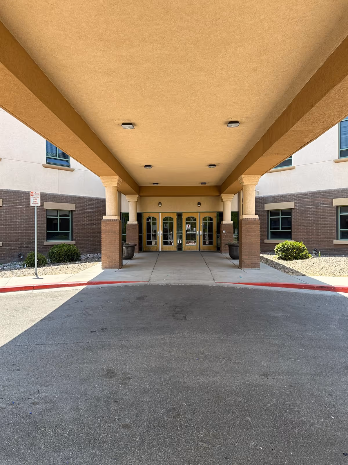 Covered entrance area of a building with beige ceiling and columns, brick lower walls, and glass double doors at the end. There are small bushes and a no parking sign on the left side near the building.