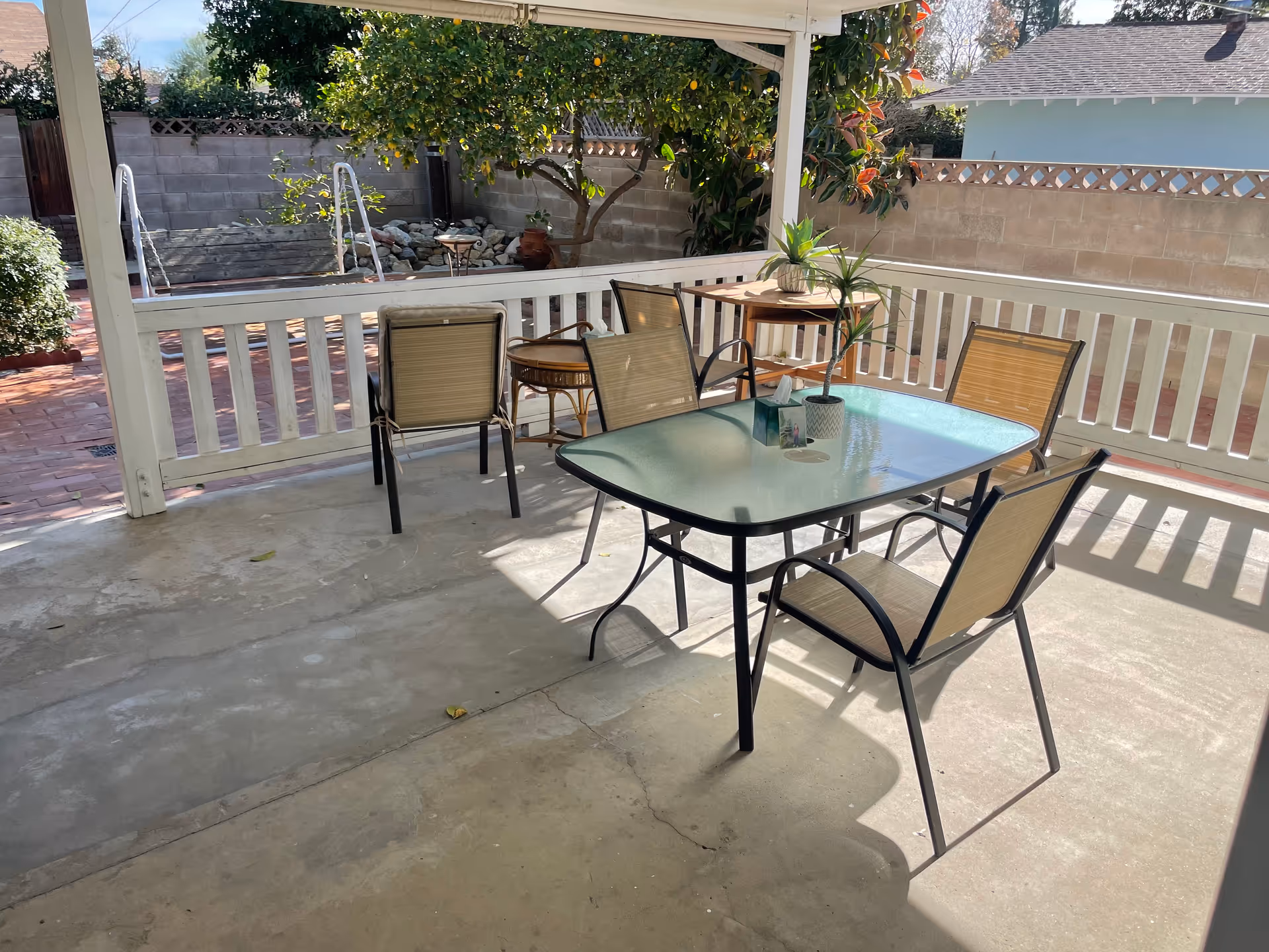 Covered outdoor patio area with a glass-top table and five beige mesh chairs. There are two small potted plants and a tissue box on the table. The patio has a white railing and overlooks a garden area with a tree, some bushes, and a small rock feature. The ground is concrete and there is sunlight casting shadows on the patio floor.