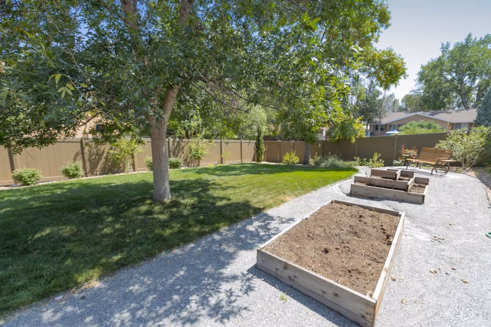 Outdoor garden area with raised wooden planting beds along a gravel path, a grassy lawn, trees, and a bench near a fence.