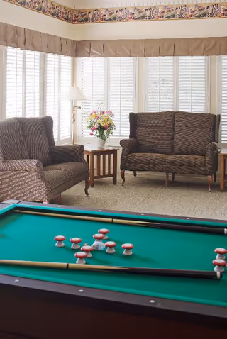 A cozy common area with a green pool table in the foreground, two brown upholstered armchairs and a loveseat arranged around a small wooden table with a floral arrangement. The room has large windows with white shutters and beige valances, and a floor lamp beside one of the chairs.