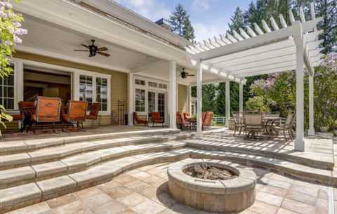 Sunlit outdoor patio with stepped stone terraces, a circular fire pit, dining sets, and a white pergola attached to a covered porch.