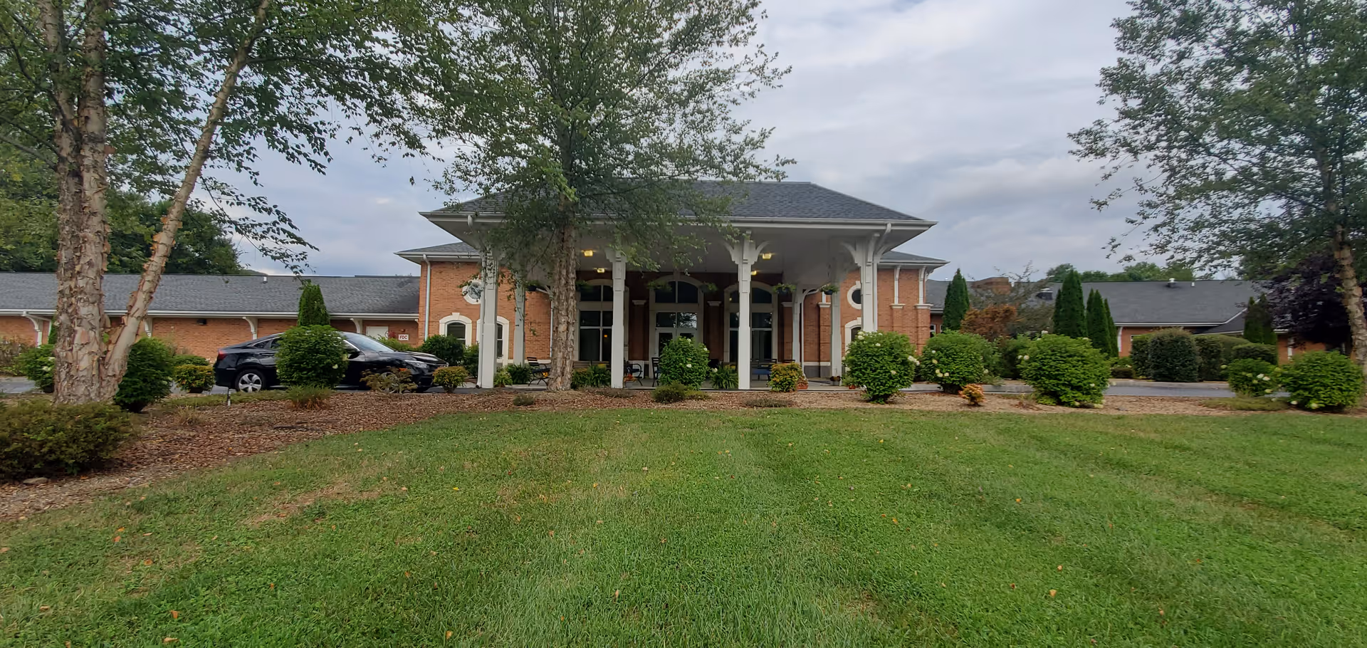 Front exterior view of a brick building with a covered entrance supported by white columns, surrounded by neatly trimmed bushes and trees, with a green lawn in the foreground and a parked black car to the left.