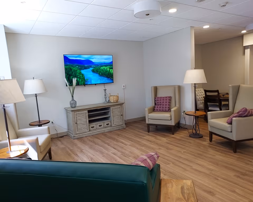 A cozy living room area with a wall-mounted flat screen TV displaying a river and forest scene. The room features light-colored walls, wood flooring, and several armchairs with purple cushions arranged around small wooden side tables with lamps. A wooden media console sits below the TV with decorative items on top.