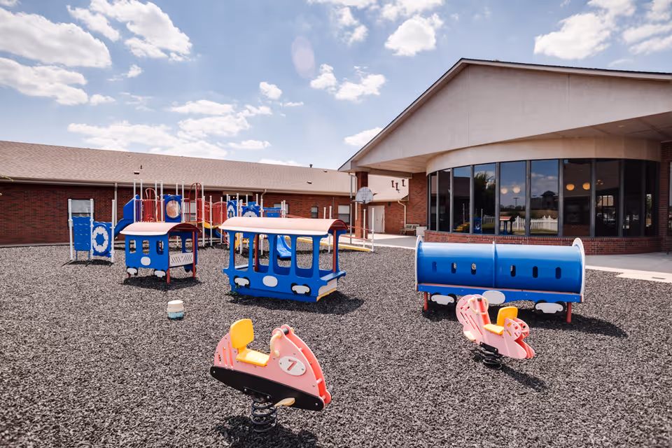 Outdoor playground area with various colorful play structures including a blue train-shaped playset, a blue tunnel, and two pink spring riders shaped like airplanes. The playground is covered with black rubber mulch and is adjacent to a brick building with large windows under a partly cloudy sky.