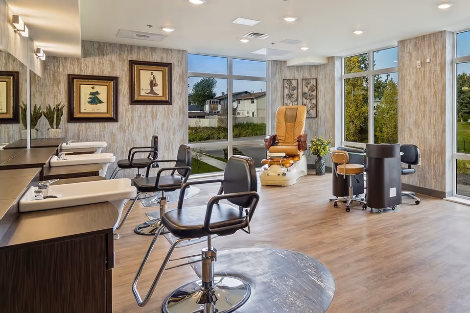A bright and modern salon area in Aspen Creek Senior Living featuring multiple black salon chairs in front of sinks and mirrors, a comfortable tan pedicure chair, and a manicure station with stools. Large windows provide natural light and views of greenery outside.