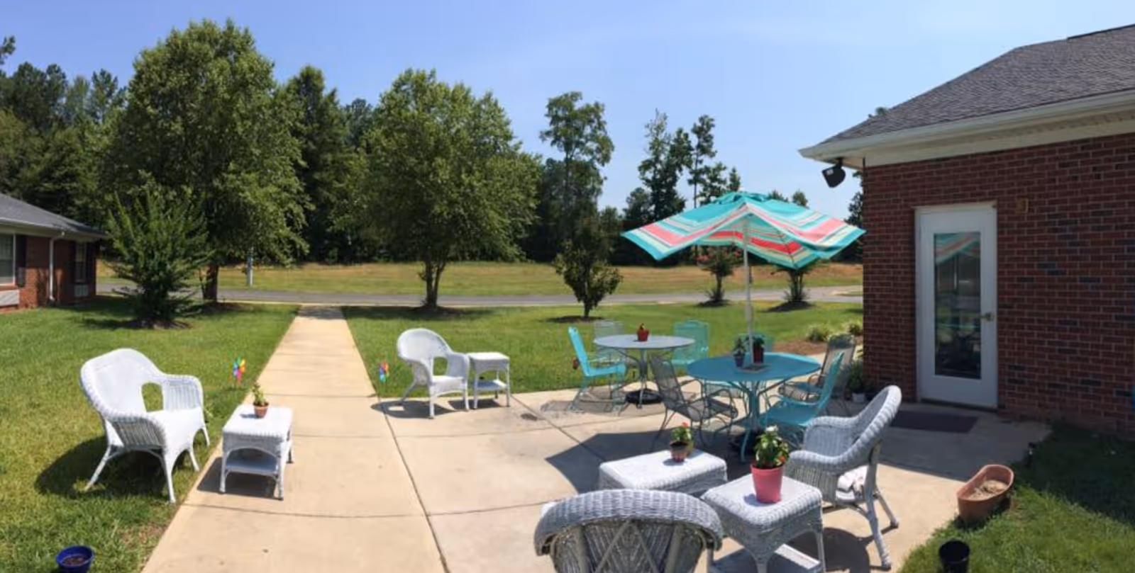 Outdoor patio area at Meadowview Terrace of Wadesboro with white wicker chairs and tables, blue metal chairs and tables with a colorful umbrella, surrounded by green grass and trees under a clear blue sky.
