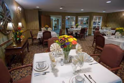 Senior living dining room with round tables, white linens, and colorful flower centerpieces.
