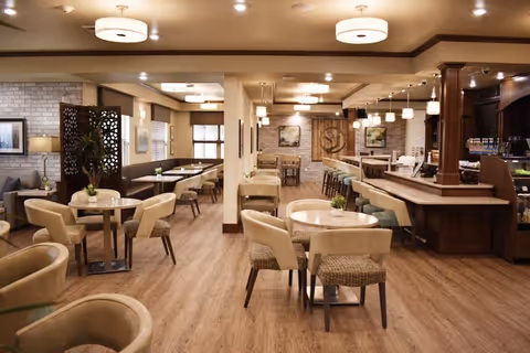 Bright, spacious dining/lounge area with round tables, upholstered chairs, booths and a service counter in a senior living facility.