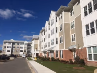 Exterior front of a multi-story beige and white independent living apartment building with a sidewalk, small lawn, and parking under a blue sky.