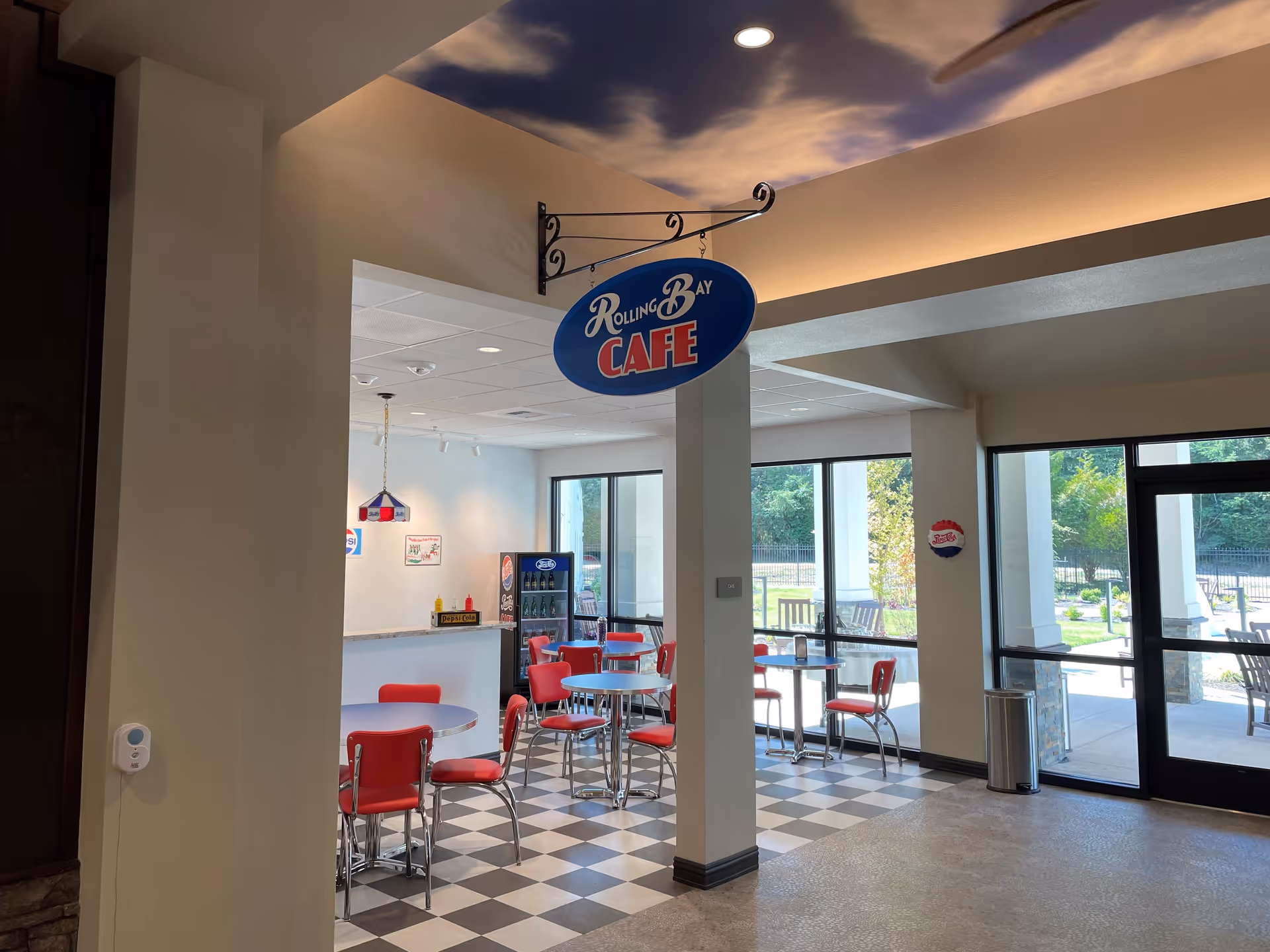 Interior view of a cafe area named Rolling Bay Cafe with red chairs and round tables on a black and white checkered floor. The ceiling has a sky mural with clouds. Large windows and glass doors provide a view of an outdoor patio with chairs and greenery.