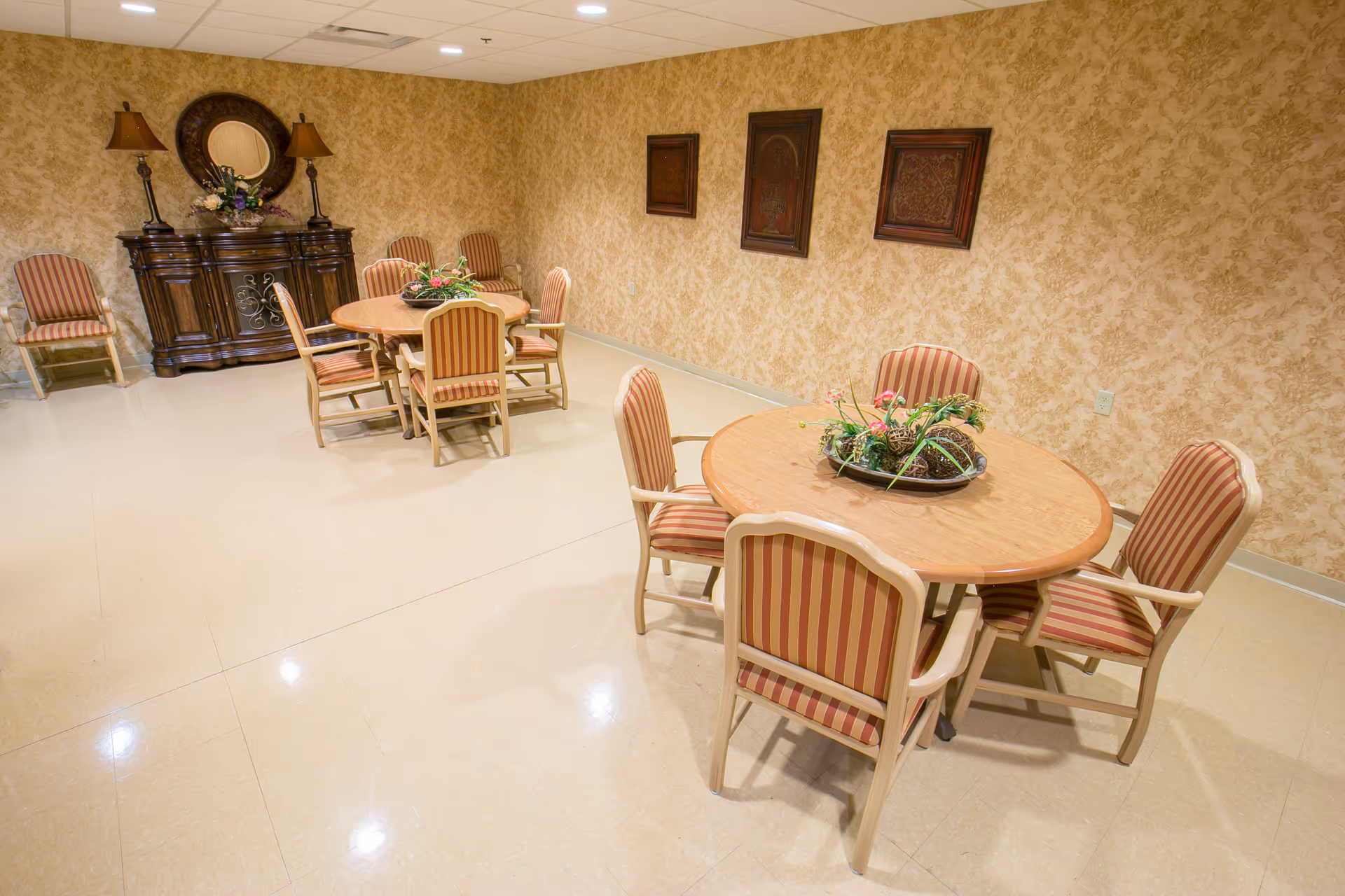 A dining room with two round wooden tables, each surrounded by four striped cushioned chairs. The room has beige patterned wallpaper, a polished tile floor, and a wooden sideboard with a round mirror and two lamps on top. There are three framed artworks on the wall.