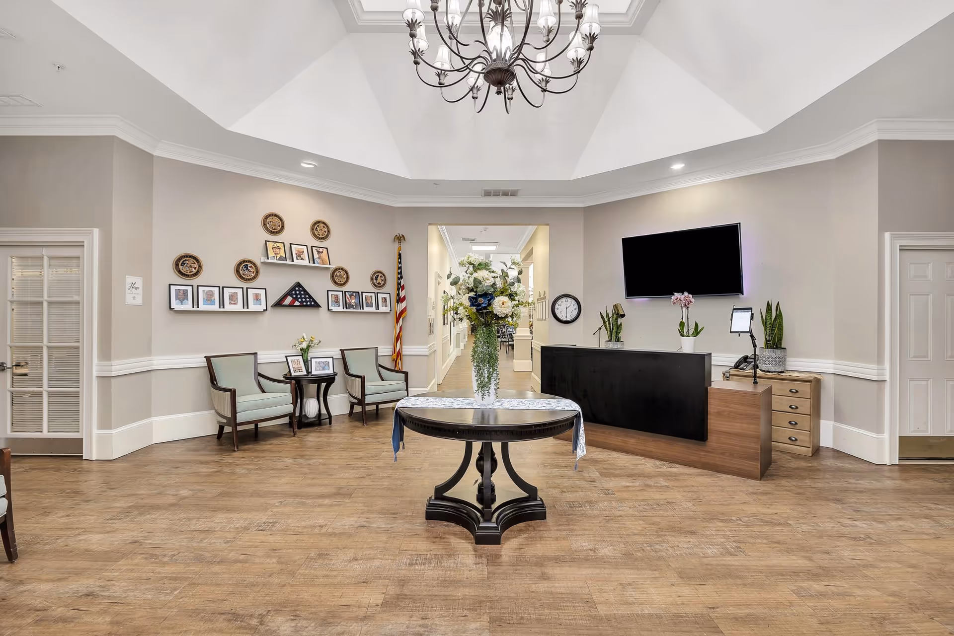 A spacious and well-lit reception area with a round table in the center holding a large floral arrangement. To the right is a reception desk with plants and a wall-mounted TV above it. On the left side, there are two chairs with a small table between them, and a wall decorated with framed photos and plaques. An American flag stands in the corner near a hallway leading further into the facility. The room has a high, white ceiling with a chandelier and wood flooring.