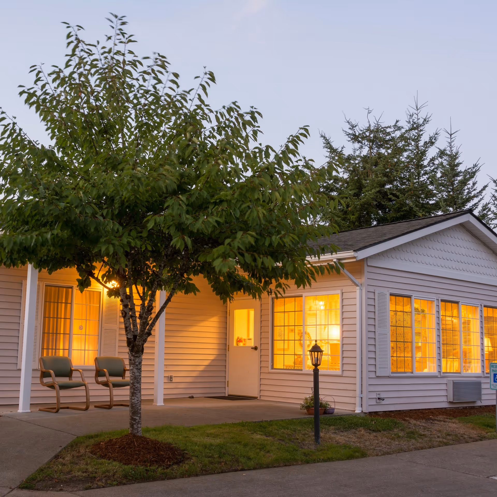 Exterior of a single-story senior living building at dusk with warm-lit windows, a front porch with chairs, and a tree in the foreground.