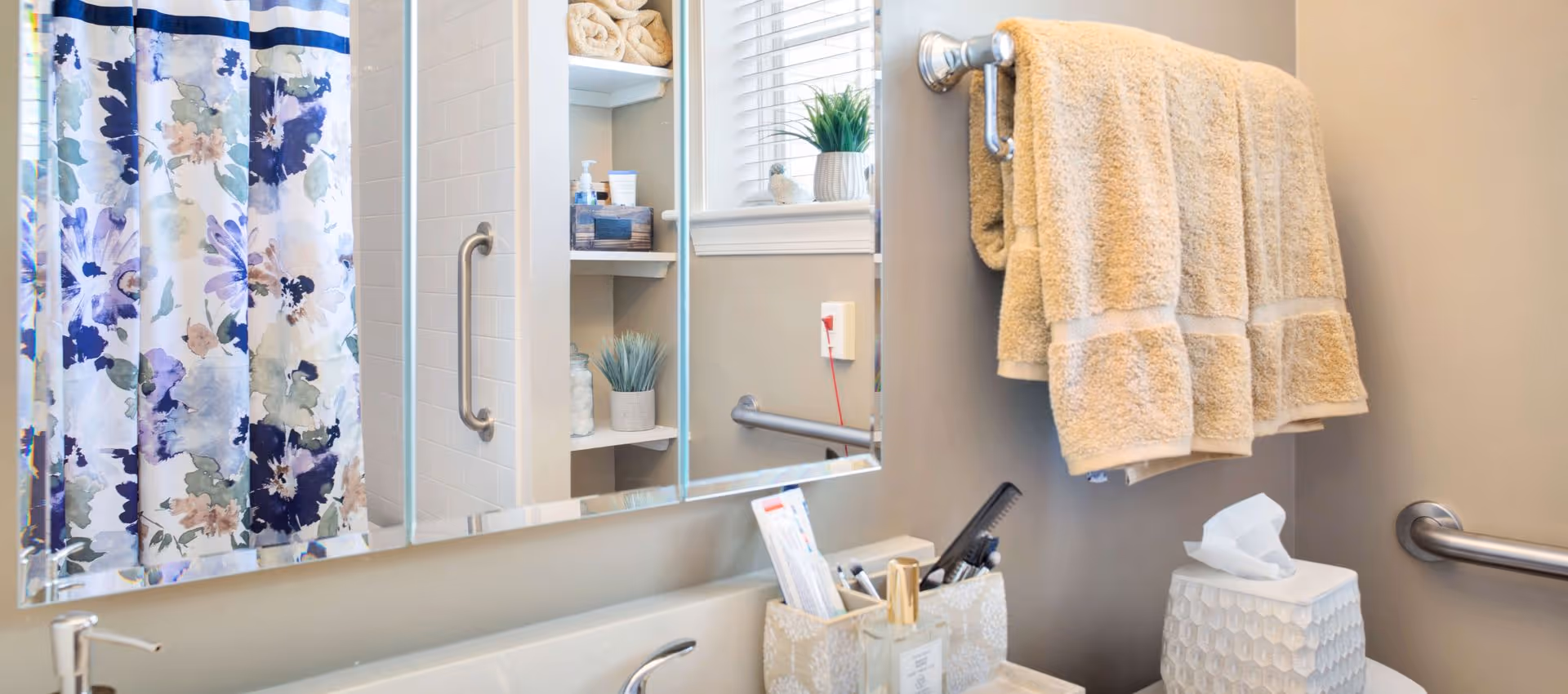 Bathroom with a floral shower curtain, vanity and mirror with toiletries, beige towels hanging on a bar, grab bars, and built-in shelves with rolled towels and decor.