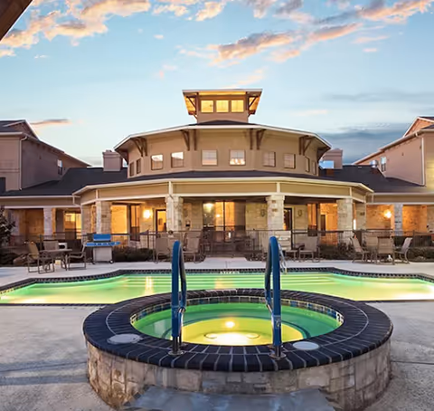Outdoor pool and illuminated hot tub in front of a multi-story senior living building at dusk.