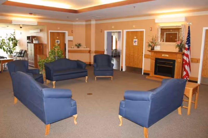A nursing home common area with four blue upholstered armchairs arranged in a circle on a carpeted floor. The room has beige walls, a fireplace with a mirror above it, an American flag, several doors, and some plants. There is also a wooden side table and a ceiling with recessed lighting.