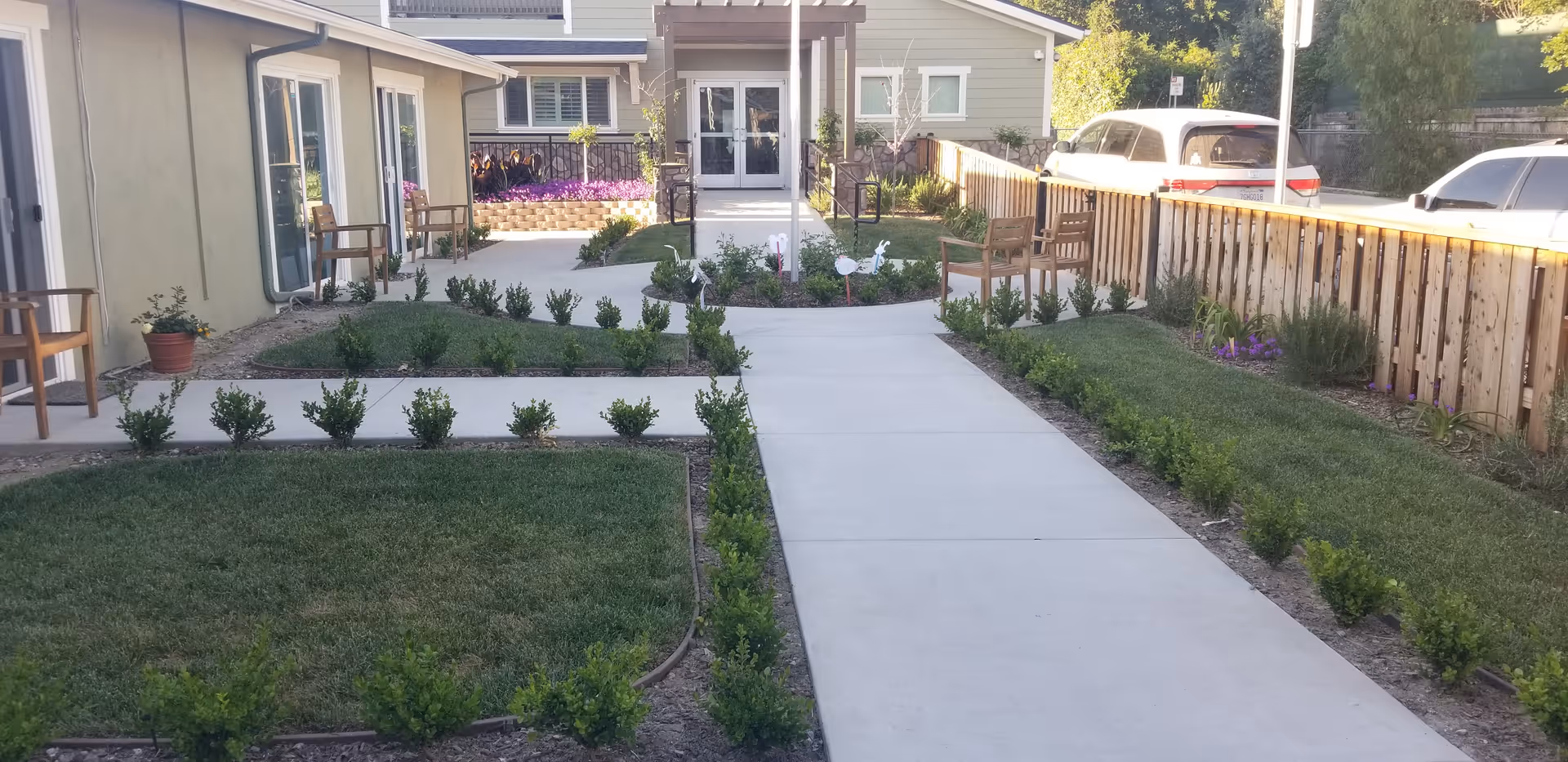 Outdoor courtyard area at Mountain Vista of Ojai featuring a concrete walkway flanked by small green bushes and grass patches. There are wooden chairs along the sides near the building walls, a wooden fence on the right, and parked cars visible beyond the fence. The buildings have light-colored walls and windows with white frames.