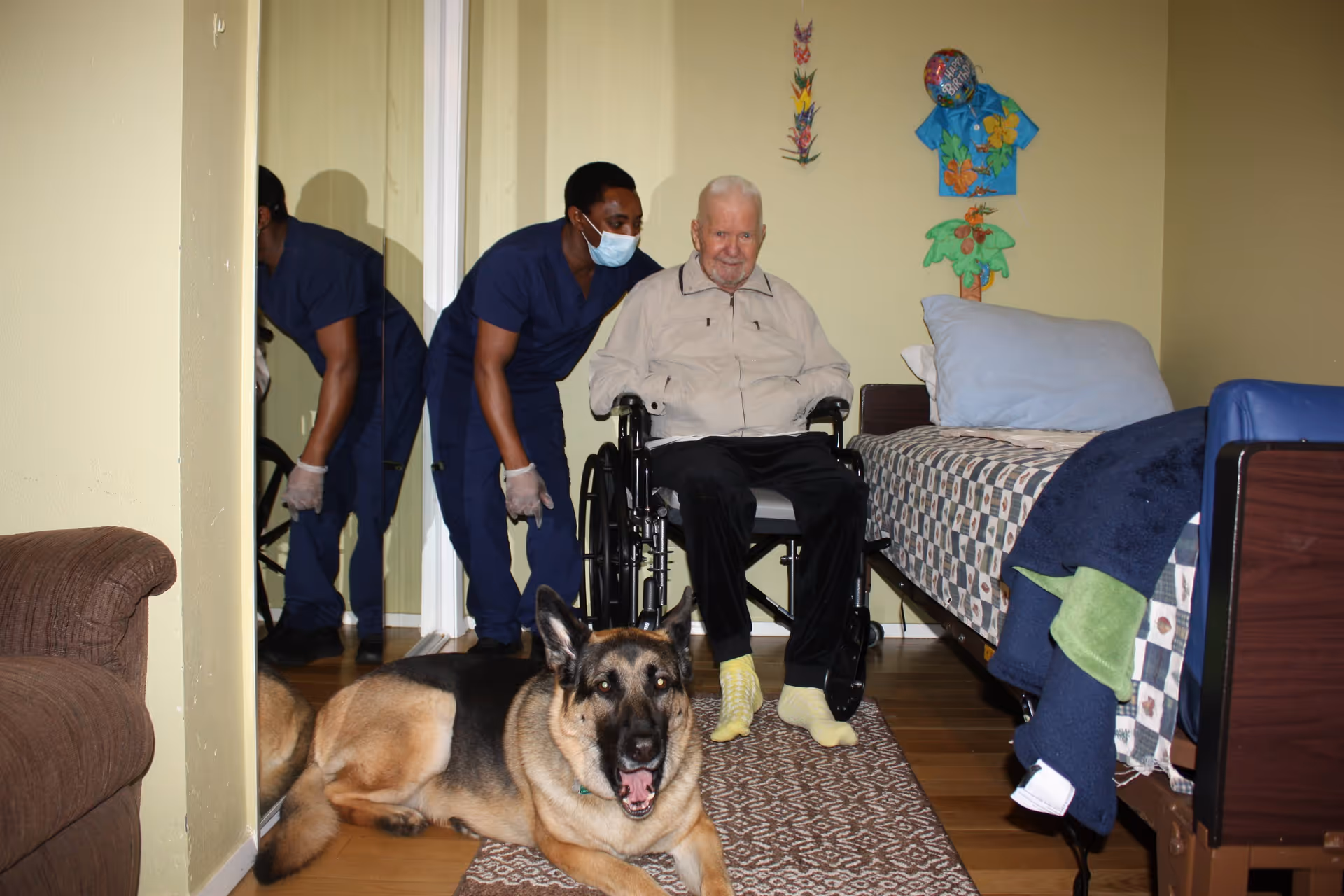 An elderly man in a wheelchair in a bedroom with a caregiver in scrubs and a large German Shepherd lying on the floor.