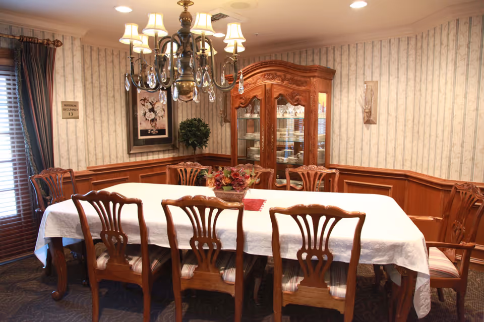 Formal dining room with a long table covered by a white tablecloth, wooden chairs, a chandelier, and a china cabinet.