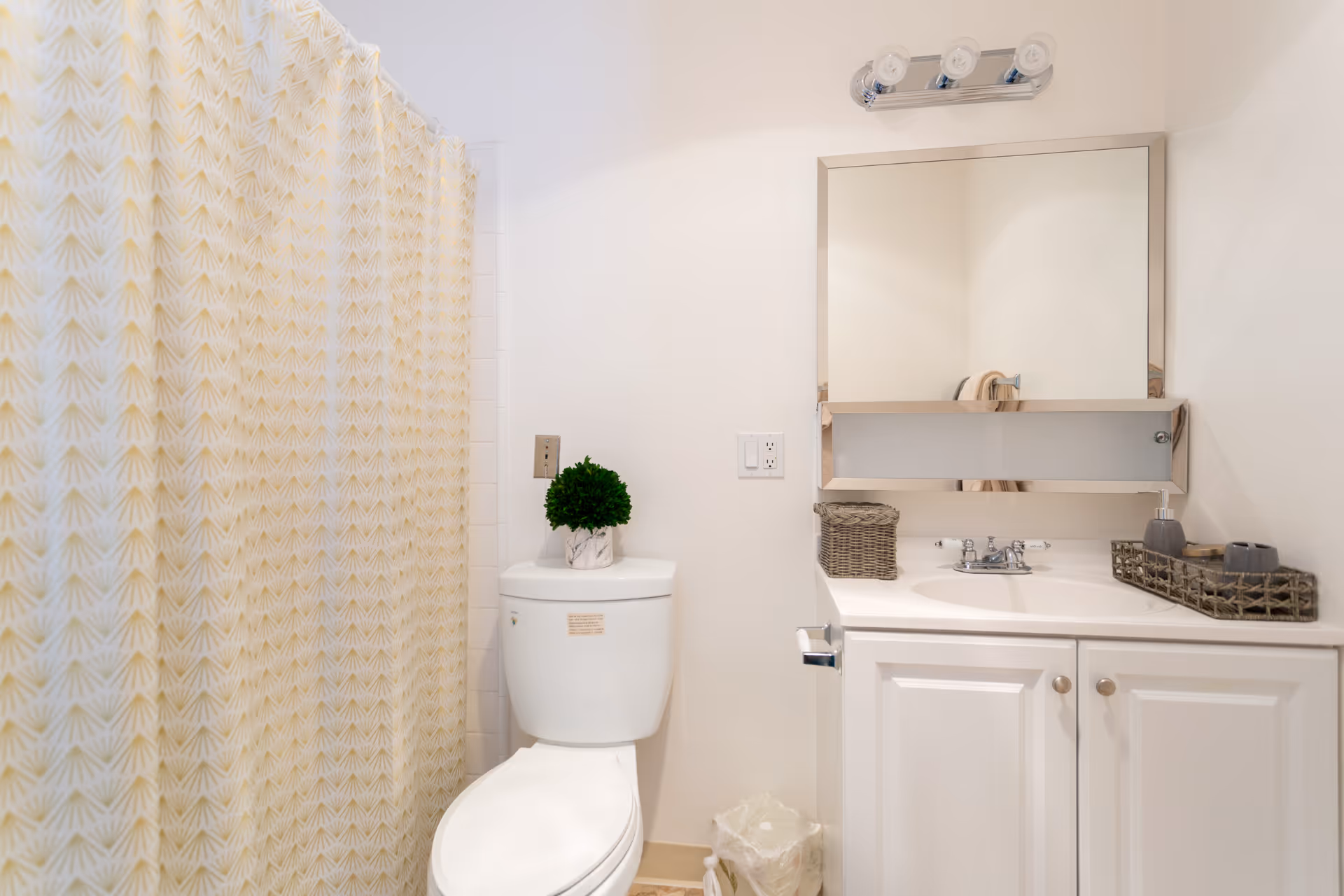 A clean bathroom with a white toilet topped with a small green potted plant, a white vanity with a sink, a large mirror above the sink, and a shower with a yellow patterned curtain.
