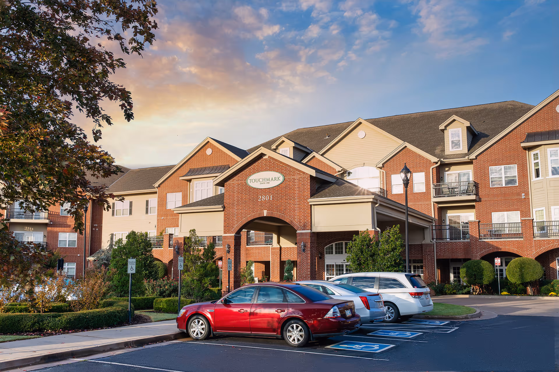 Exterior view of Touchmark at Coffee Creek, a multi-story senior living facility with brick and beige siding, featuring a covered entrance, several parked cars in front, and landscaped greenery under a partly cloudy sky at sunset.