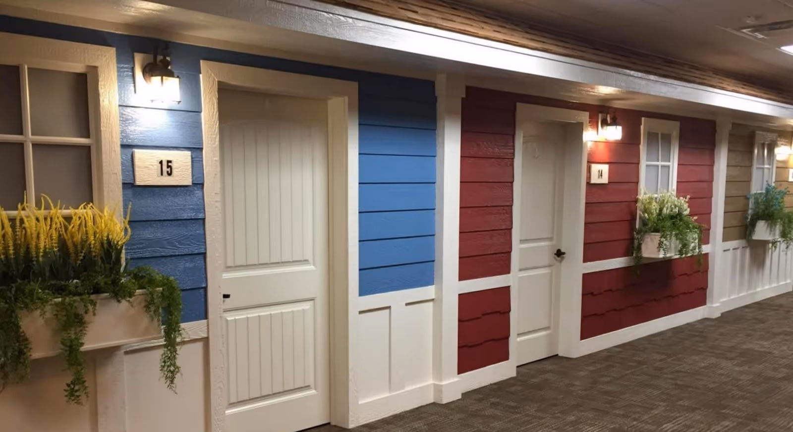 Indoor hallway with multiple doors painted in different colors including blue, red, and beige. Each door has a number plaque and a window box with green plants and flowers beneath the windows. The hallway is carpeted and well-lit with wall-mounted lights above each door.