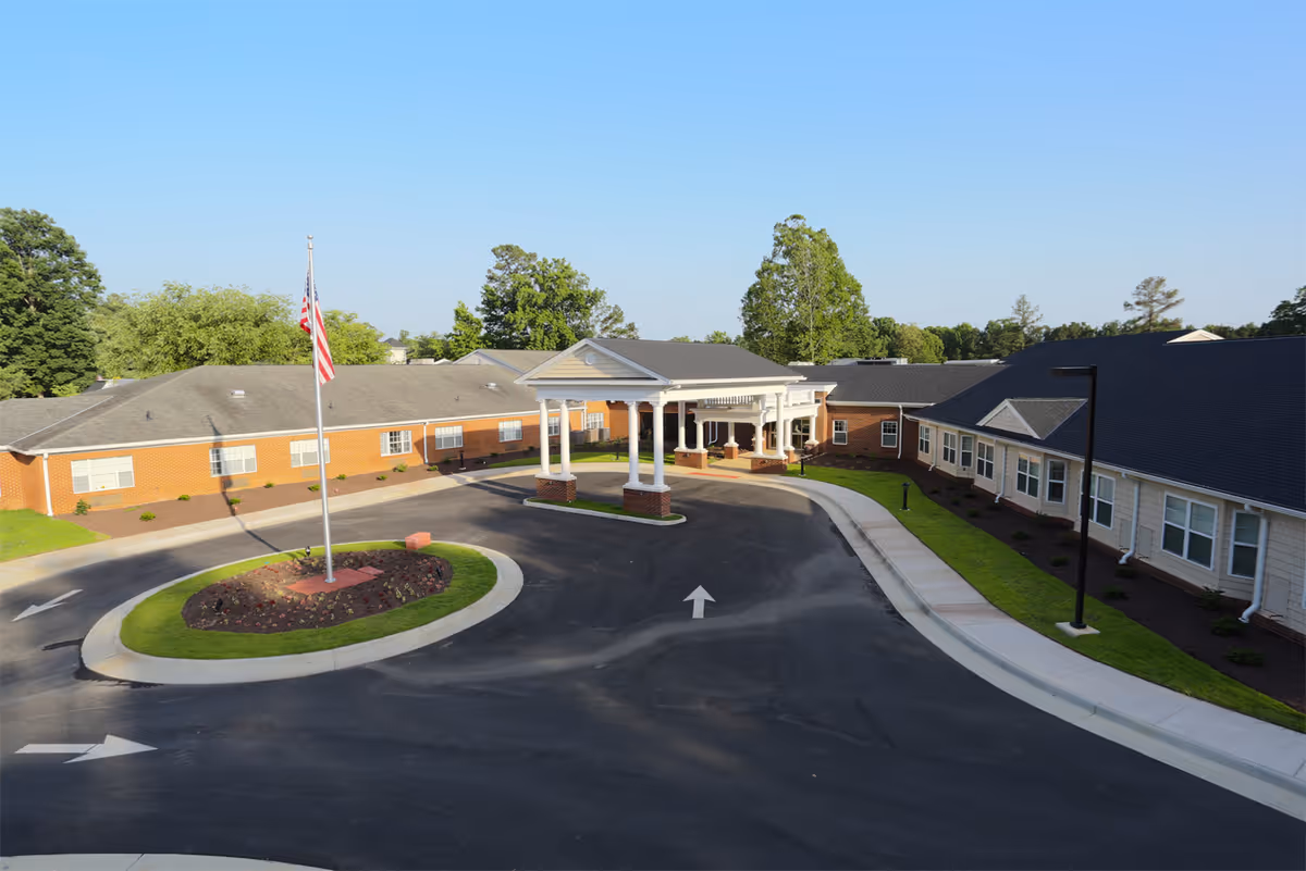 Front exterior view of Brandermill Woods Retirement facility showing a circular driveway with a flagpole in the center, a covered entrance with white columns, and single-story brick and siding buildings surrounded by greenery under a clear blue sky.