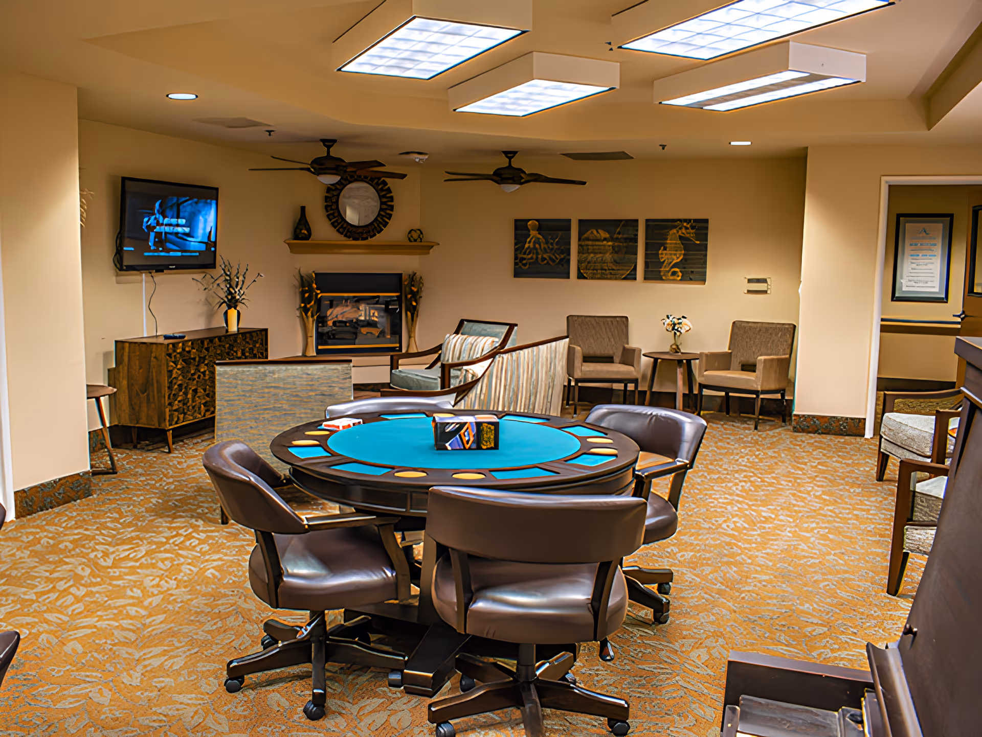 A cozy common area in a senior living facility featuring a round card table with six brown leather swivel chairs. The room has a patterned carpet, a wall-mounted TV, a fireplace with decorative vases, and several armchairs and small tables arranged around the space. Ceiling fans and fluorescent lights are visible on the ceiling. Wall art with sea creatures decorates the back wall.