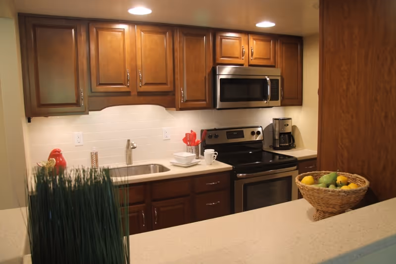 Modern kitchen with wooden cabinets, stainless steel stove and microwave, sink, and a fruit basket on the counter.